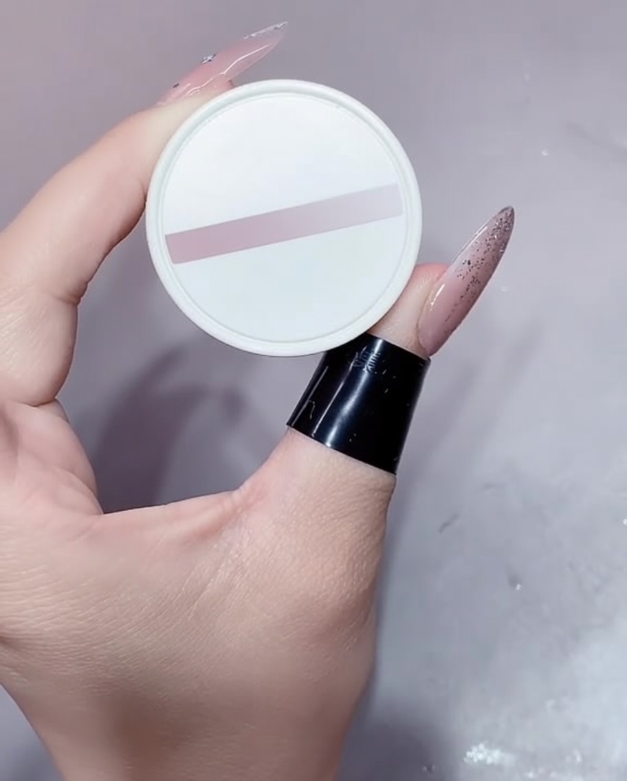 A small white pot of nude gel polish being held with a manicured hand.