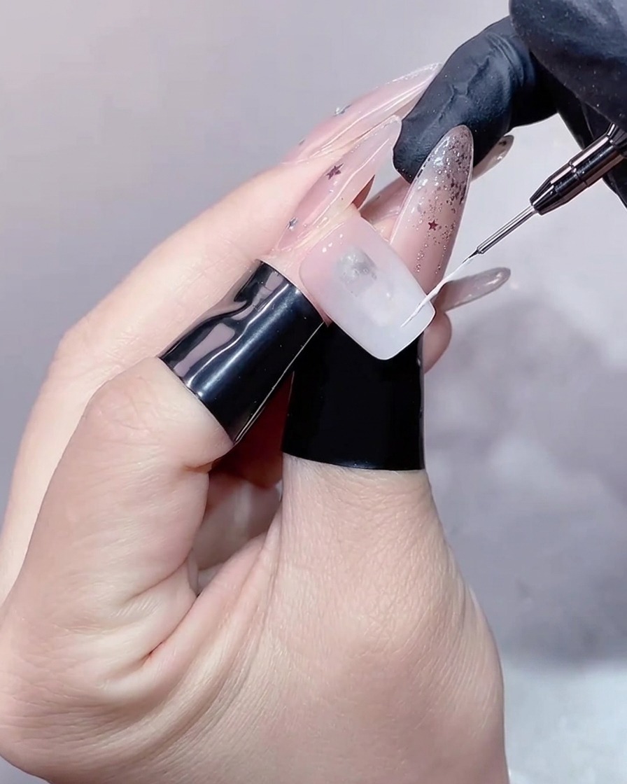 A close-up view of a thin liner brush drawing a delicate white line across the tip of a nail to create a French outline.