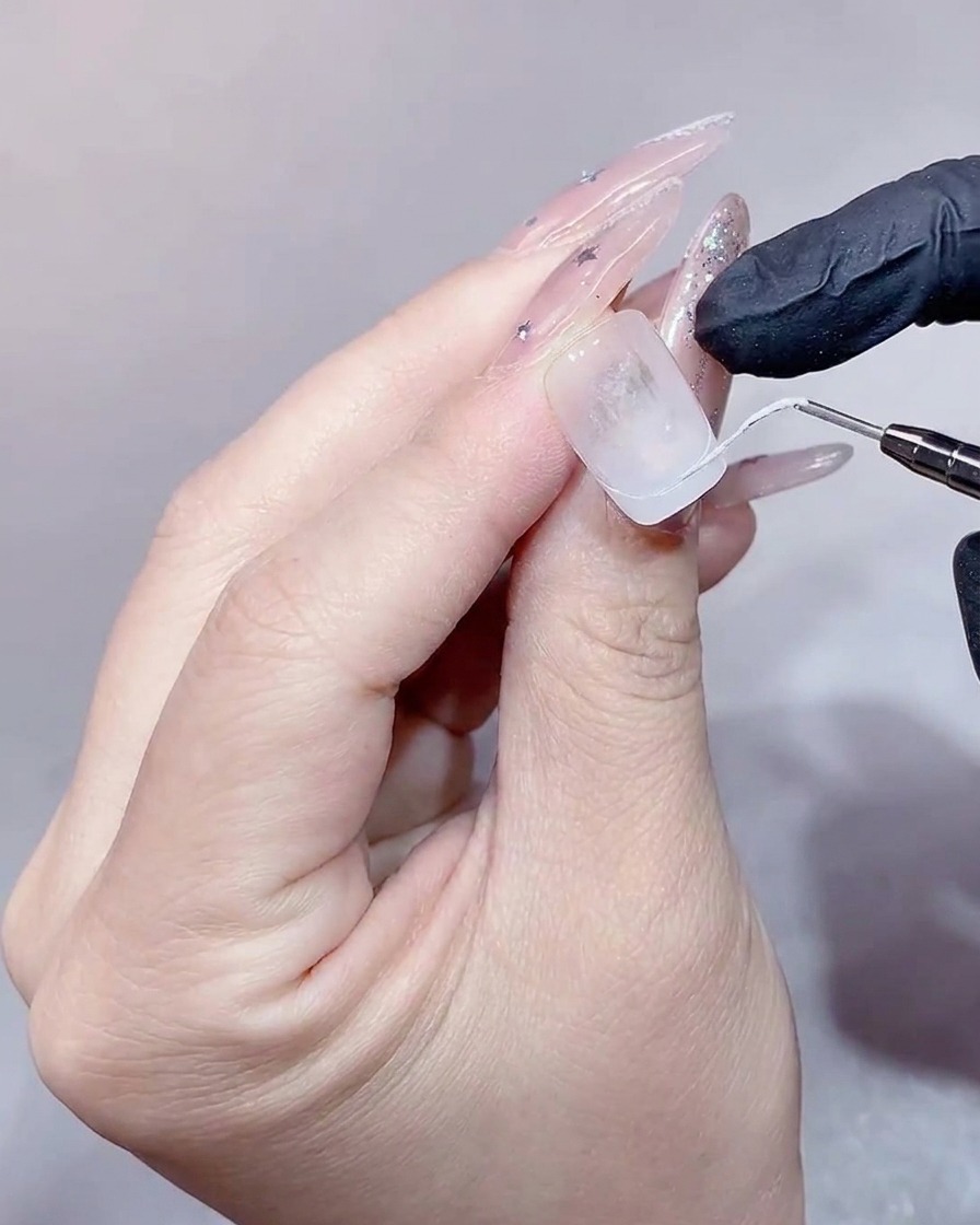 A close-up view of a nail technician using a flat brush to fill in a white French tip on a clear nail extension.