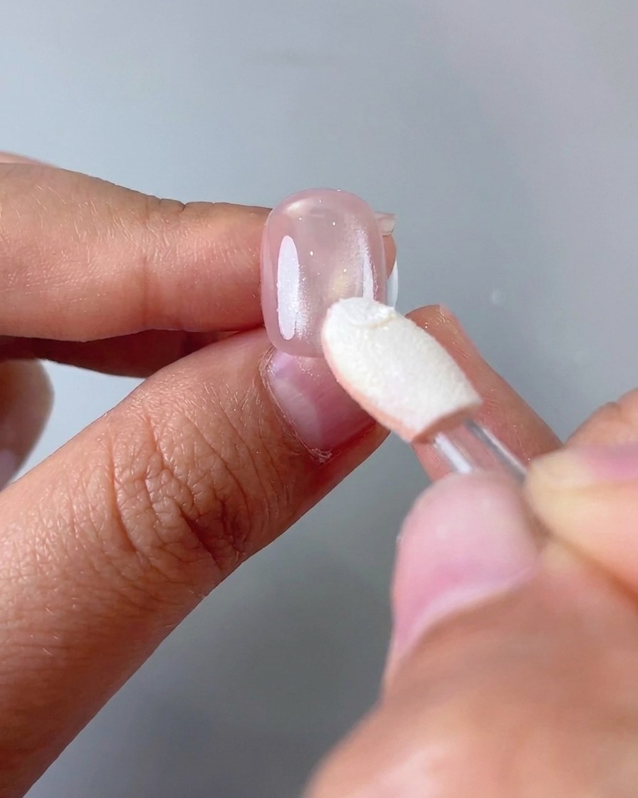 An eyeshadow sponge applicator rubbing silver mirror powder onto the bottom two-thirds of a pink nail.