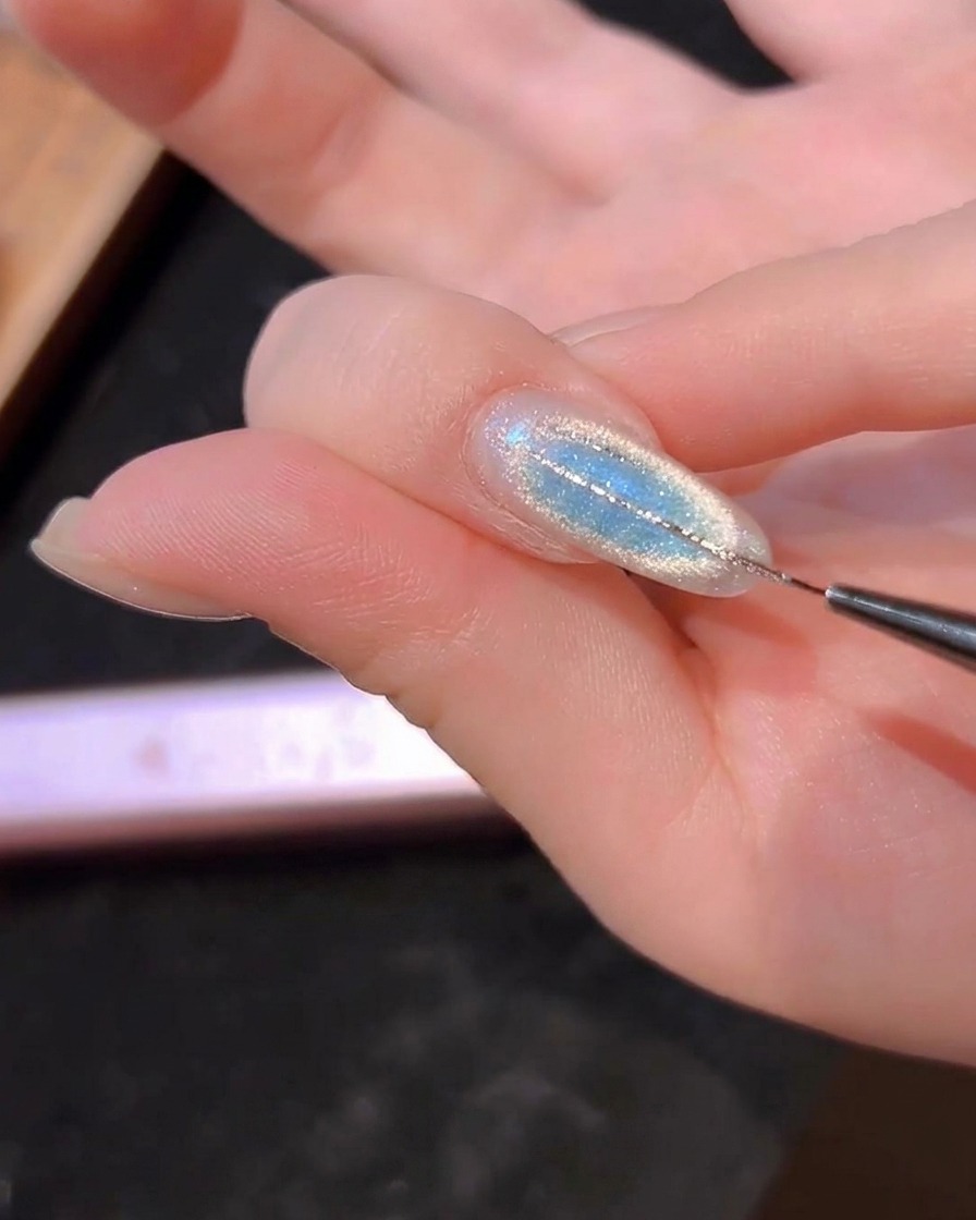 A fine liner brush drawing thin silver lines on a blue cat-eye nail.