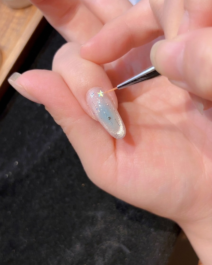 A fine brush placing a holographic star glitter onto the wet gel of a manicured nail.