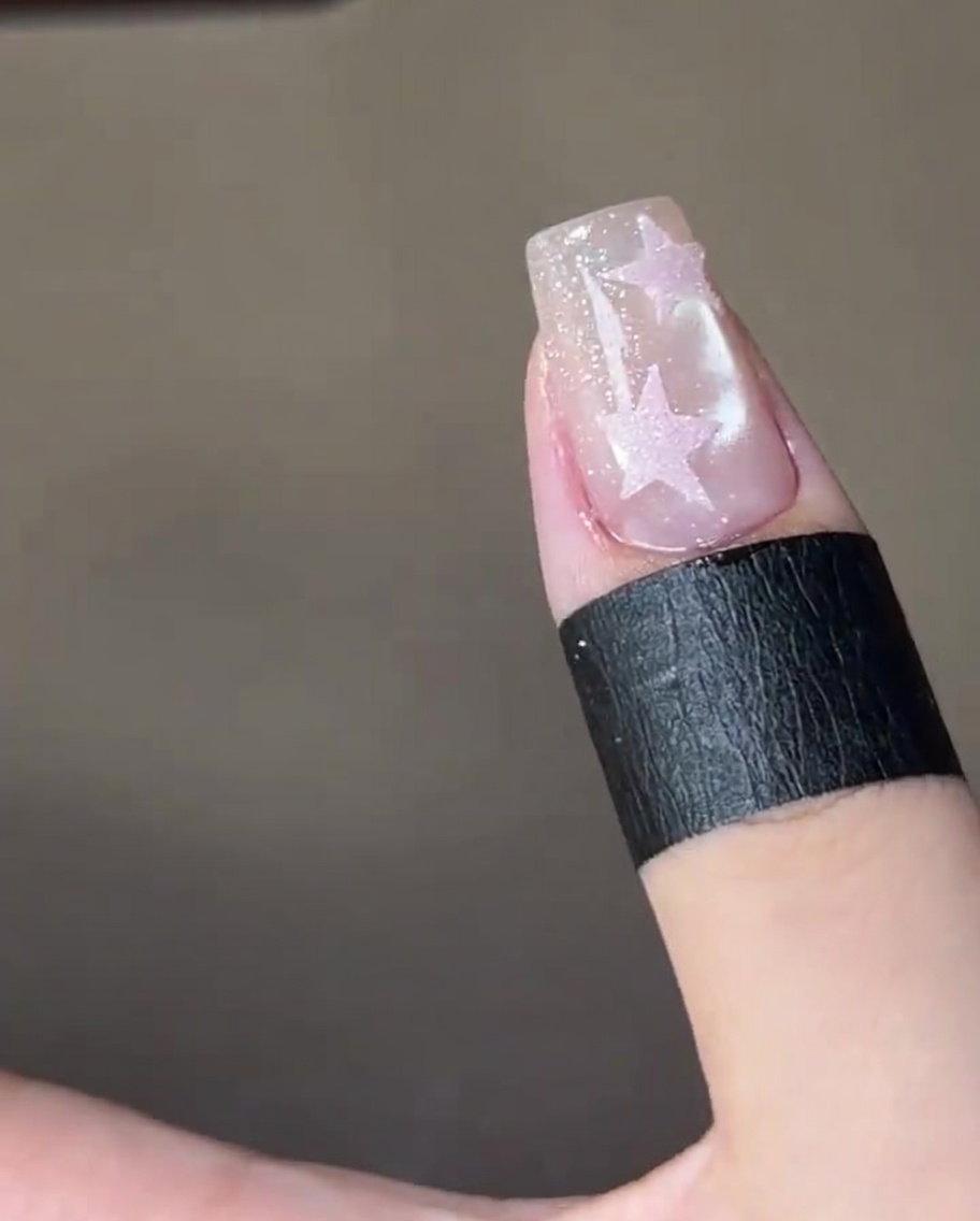 A close-up view of a brush applying a glossy clear top coat over a pink star painted on a shimmering nail.