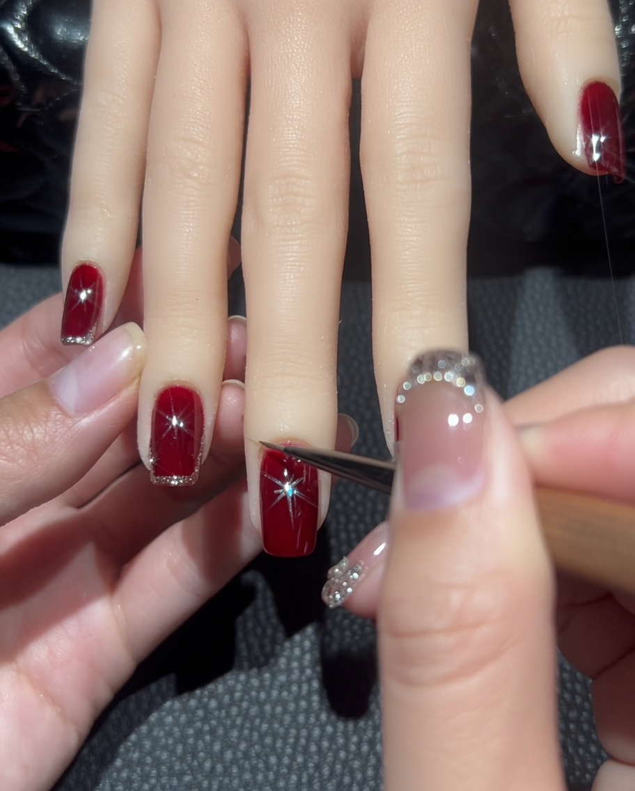 A close-up of a liner brush tracing a thin line of rose-gold glitter around the edge of a deep red nail.