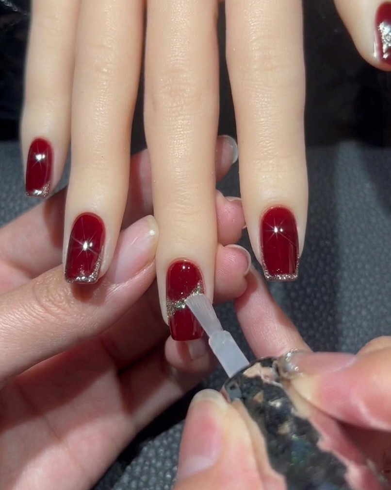 A brush applying a clear top coat to a red nail with a glitter design, carefully navigating around a central rhinestone.