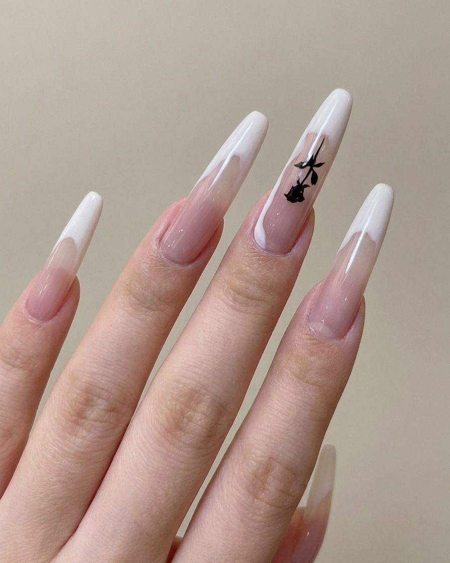 A close-up of a nail brush drawing a thin, black branch pattern on a pre-painted French tip nail.
