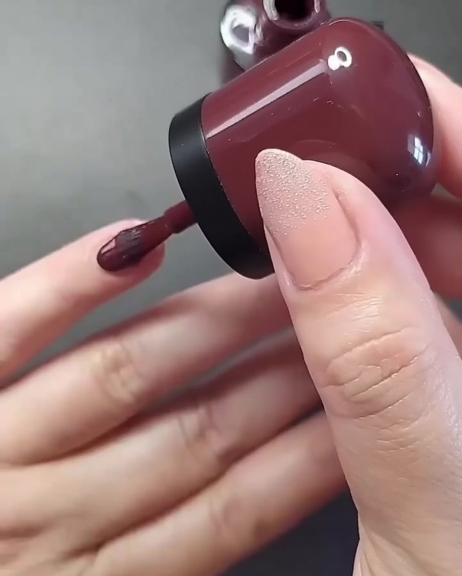 A close-up of a hand with a nail brush applying deep burgundy polish to the pinky fingernail.