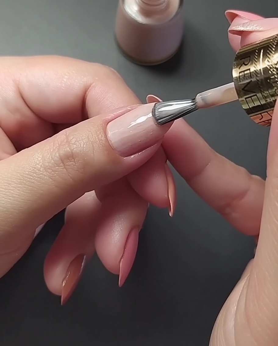 A close-up view showing a thin metallic gold line being painted vertically down the center of a light nude thumb nail.
