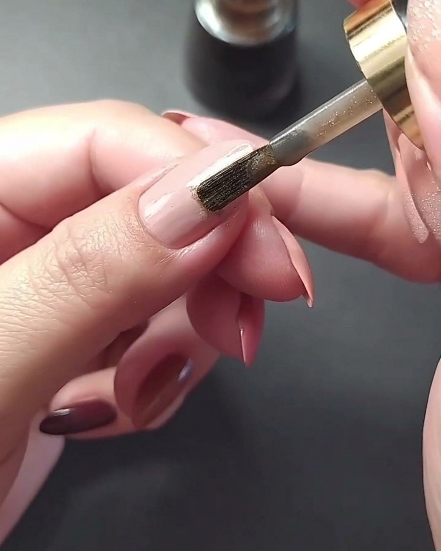 A macro shot showing a thin metallic gold line being painted vertically down the center of a mauve-colored fingernail.