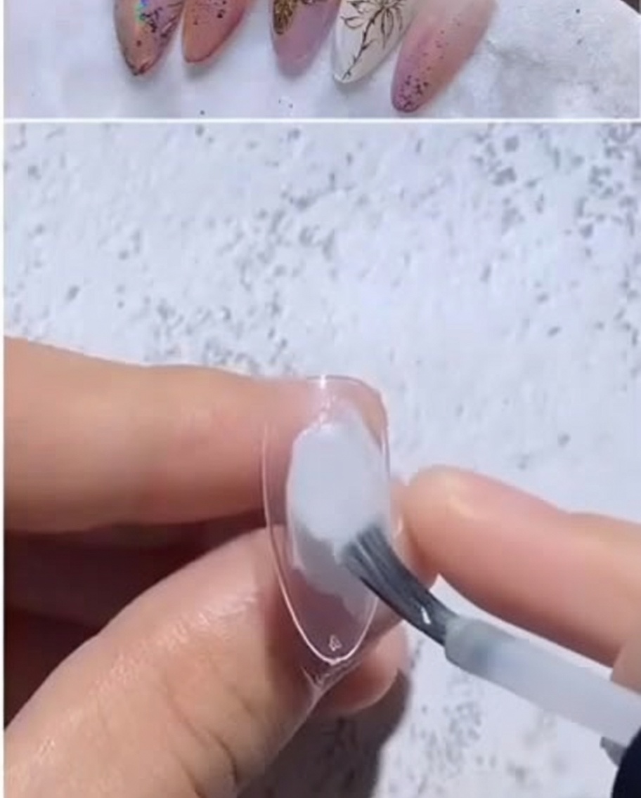 A close-up of a clear almond nail tip receiving a dot of milky white gel polish from a brush.