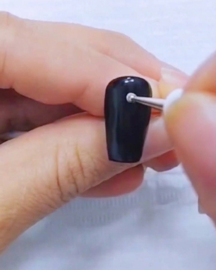A close-up of a dotting tool placing a small white dot on a solid black nail.
