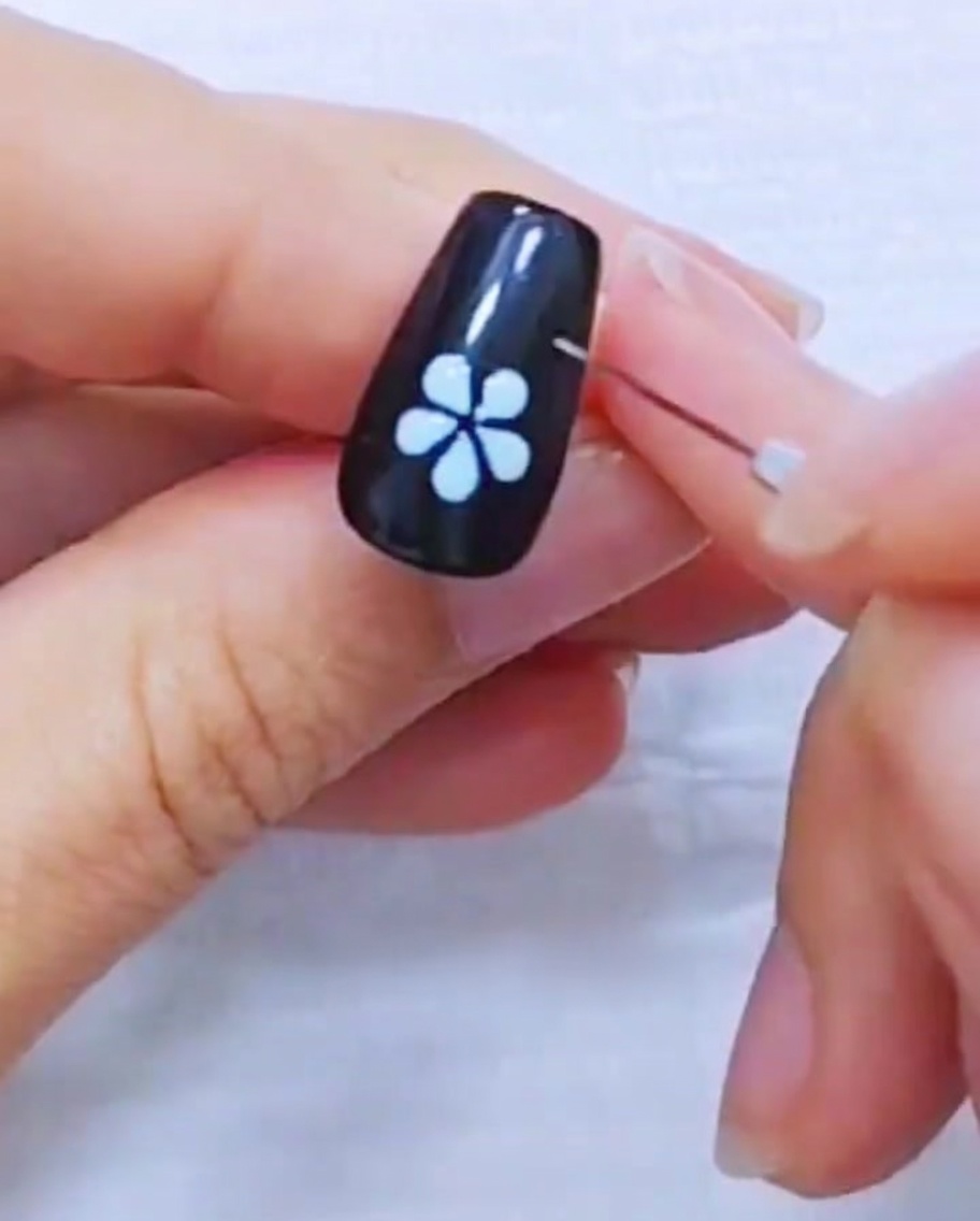 A close-up of a liner brush refining the edges of a white five-petal flower design on a black nail.
