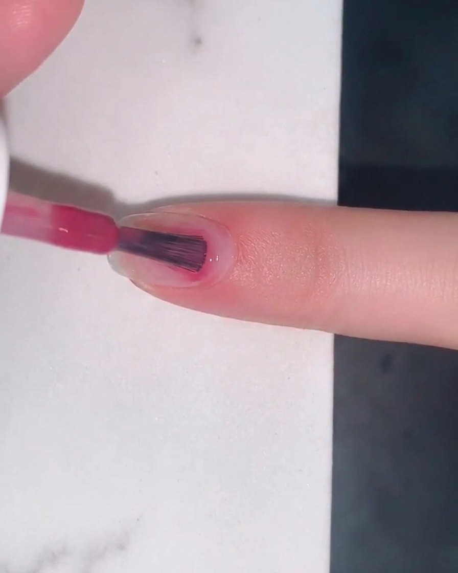 A brush applying a sheer layer of berry-pink jelly gel polish to a natural fingernail.