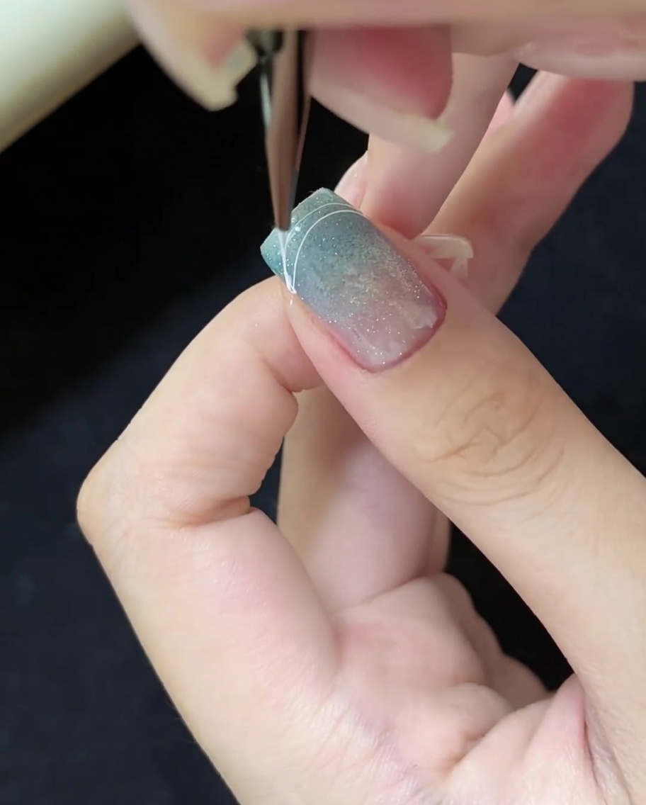 A close-up of a nail brush drawing a thin white curved line on the right corner of a blue cat-eye nail tip.