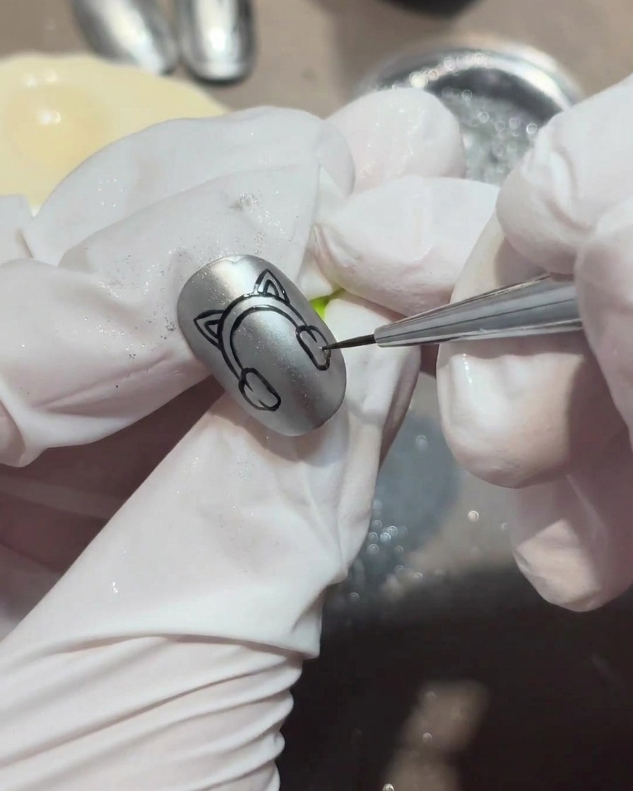 A close-up of a gloved hand using a fine liner brush to paint black kitten ear shapes onto a silver chrome nail.