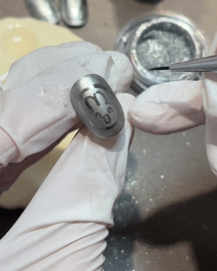 A close-up of a liner brush applying a small, clear gel bead onto a matte gray nail to form a water droplet.