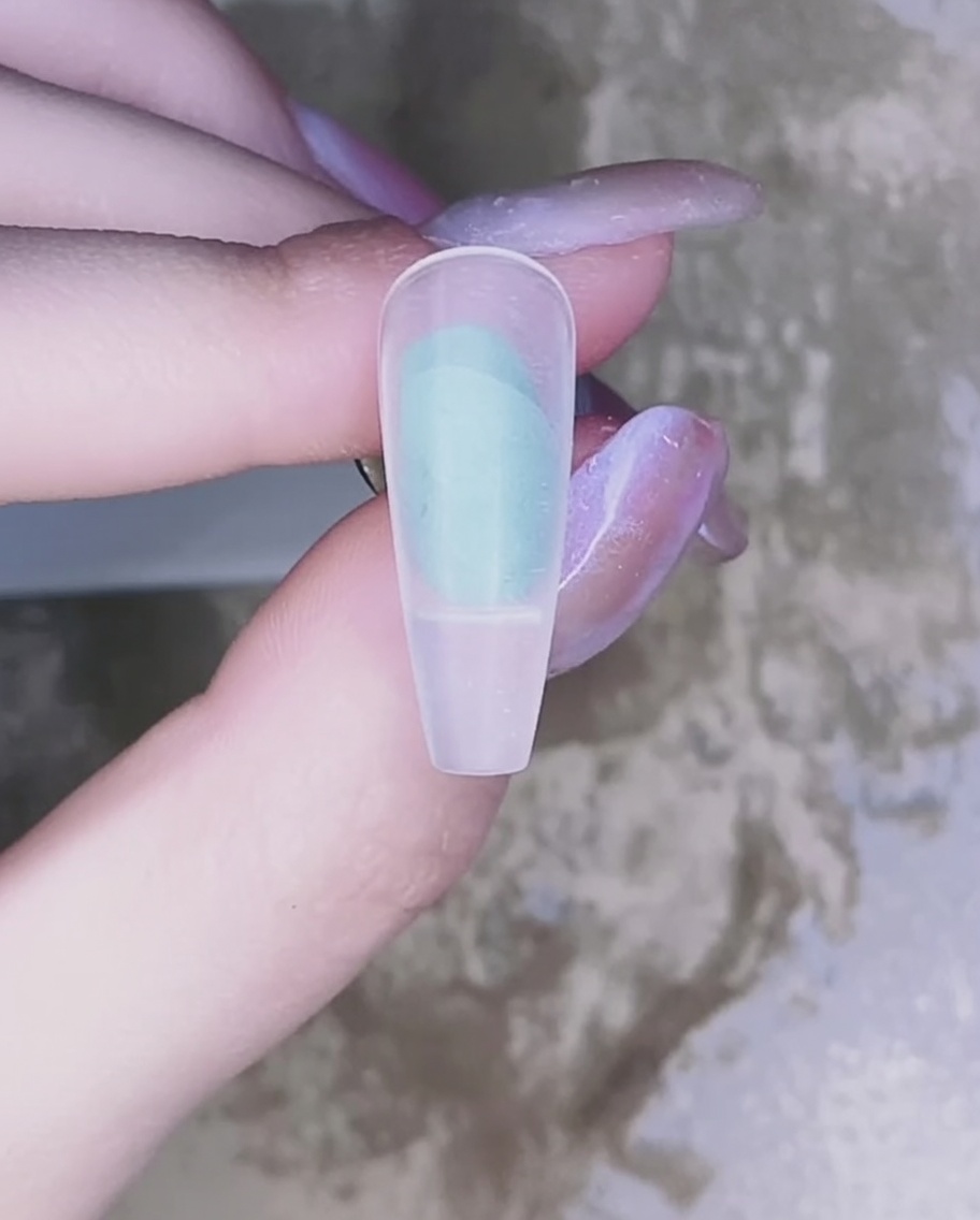 A close-up of a brush applying sheer pink gel polish onto a natural nail surface.