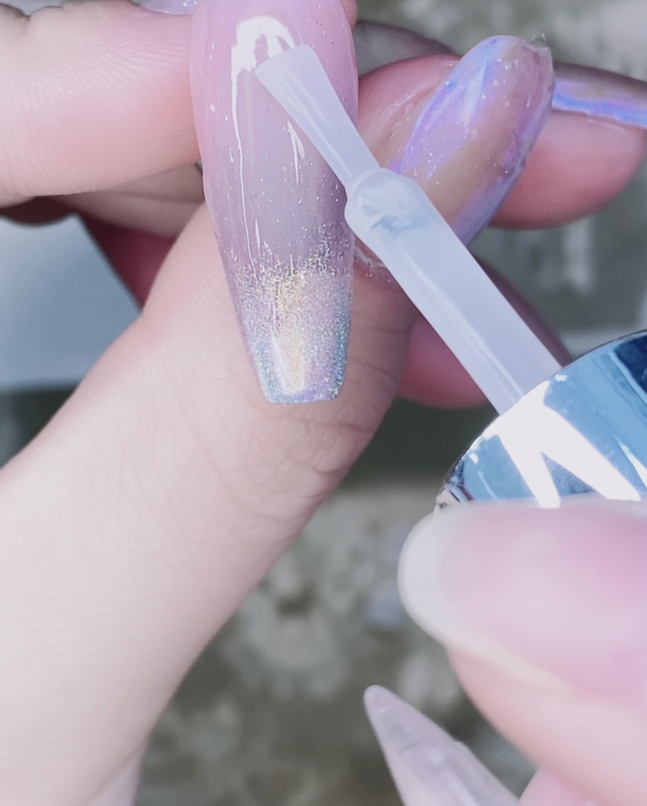 A nail brush applying clear top coat over the chrome-powdered gradient on the nail.