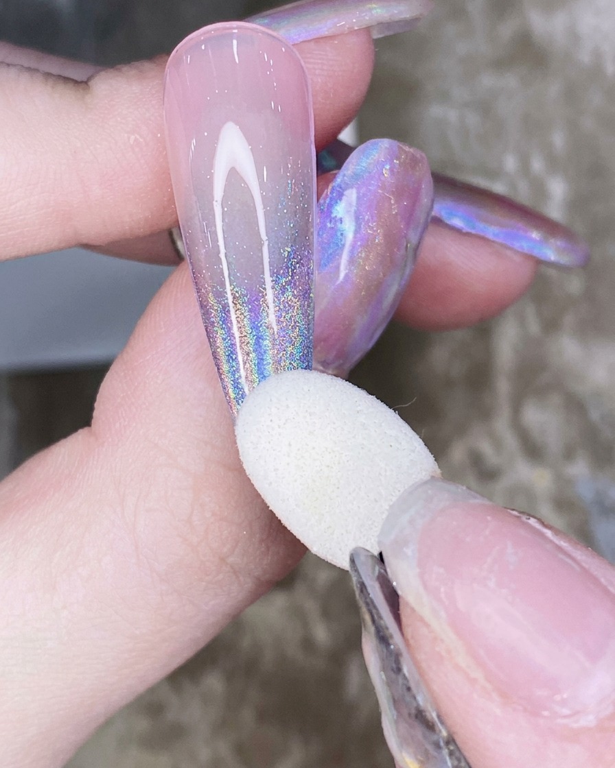 A close-up of a finger holding a nail with a sponge applicator brushing aurora chrome powder in an upward gradient motion.