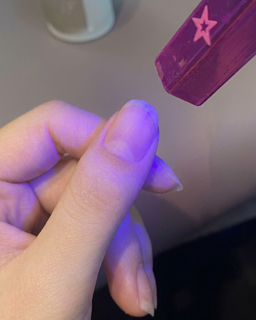 A hand placing a fingernail under a small, handheld purple UV lamp to cure the base coat.
