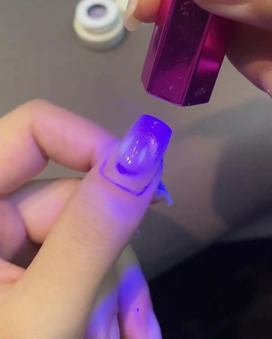 A purple cat eye gel nail being cured under a small handheld purple UV lamp.
