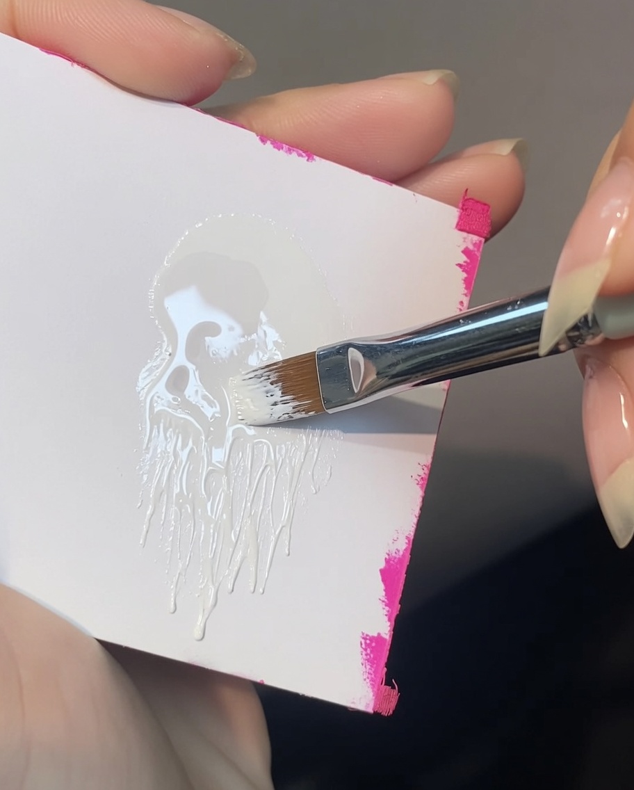 An angled nail art brush picking up white painting gel from a palette, loading only one corner of the bristles.