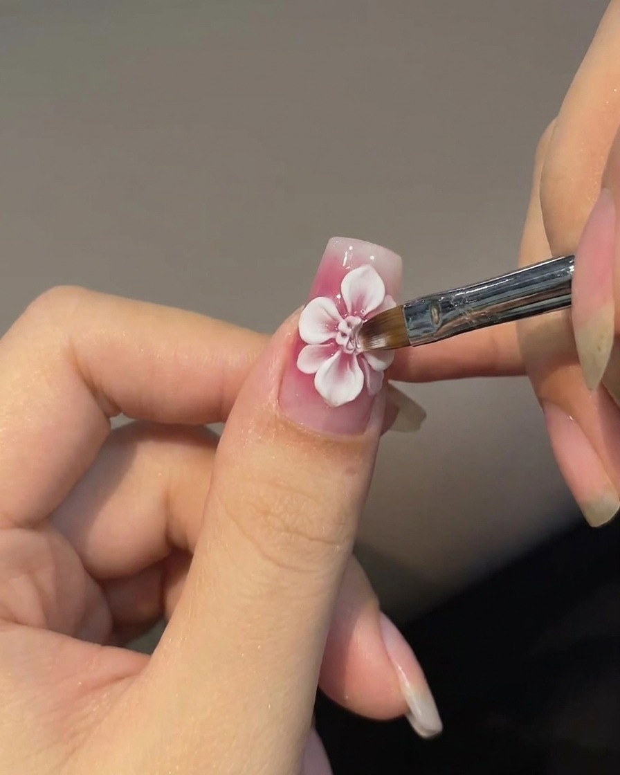 An angled nail art brush painting faded white outer petals around a red rose center on a pink nail.