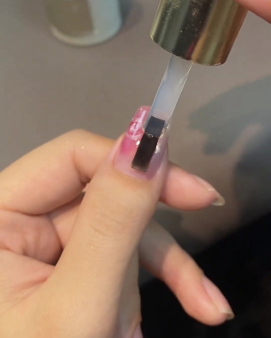 A brush applying a clear top coat over a painted white and pink rose nail design.