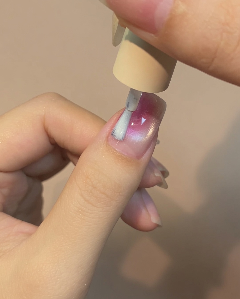 A brush applying a thin layer of clear top coat over a pink and white rose cat eye nail design.