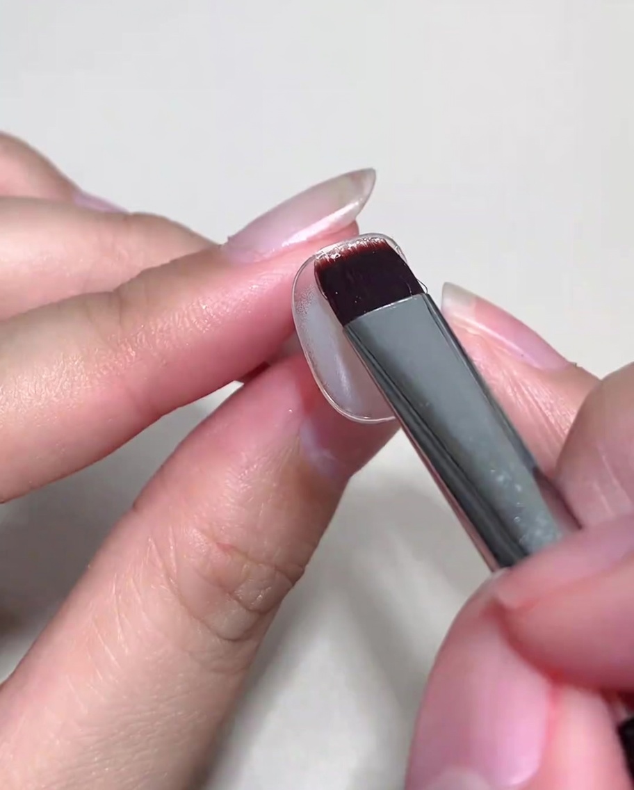 A brush applying a coat of nude cat eye gel over a cured base-coated nail.