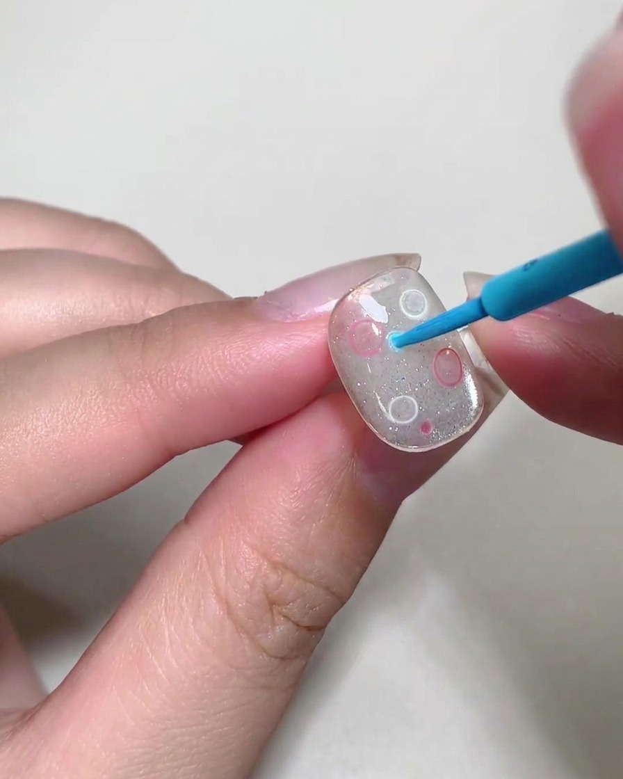 A dotting tool placing small, colorful liquid bubbles onto a nail surface.