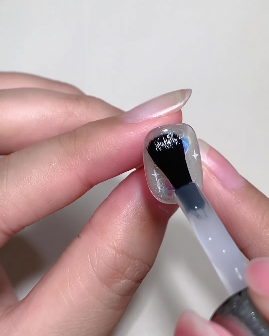 A nail brush applying a clear builder gel over a decorated nail to level the surface.