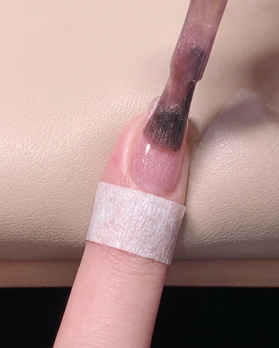 A top-down view showing a brush smoothing out the pink gel polish over the nail plate for an even finish.