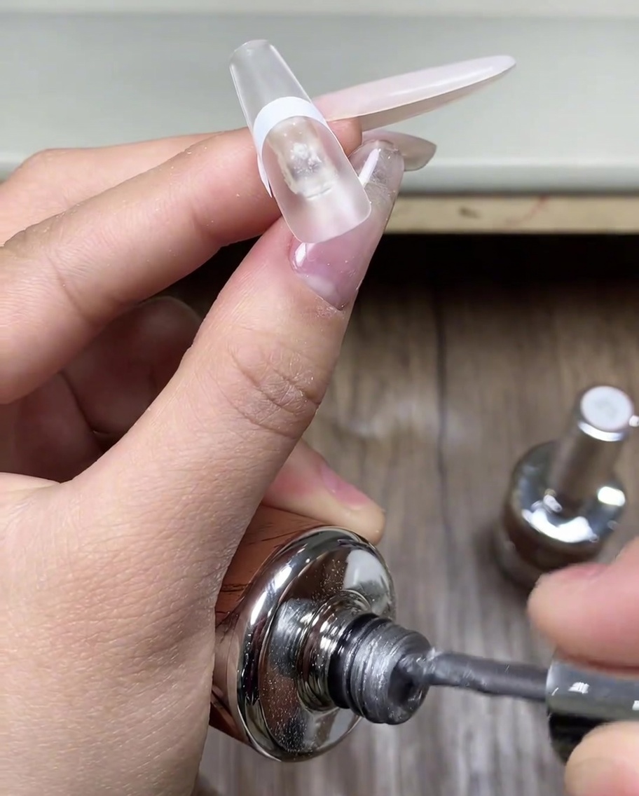 A brush applying a coat of grey cat eye gel onto the tip of a fingernail protected by a sticker.