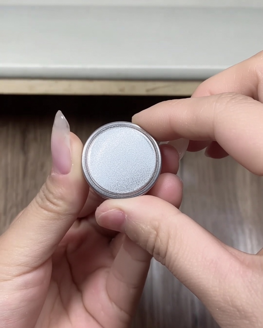 Close-up of hands holding a small, round container of silver-colored chrome powder, with the lid just removed.