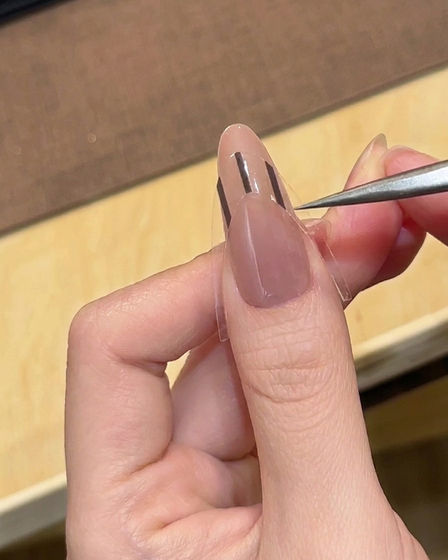 A pair of tweezers placing a black French tip guide sticker onto a nude-colored almond nail.