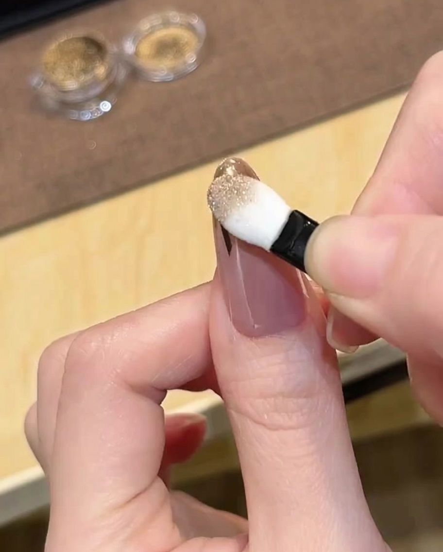 A sponge eyeshadow applicator rubbing gold chrome powder onto the tip of a French-taped nail.