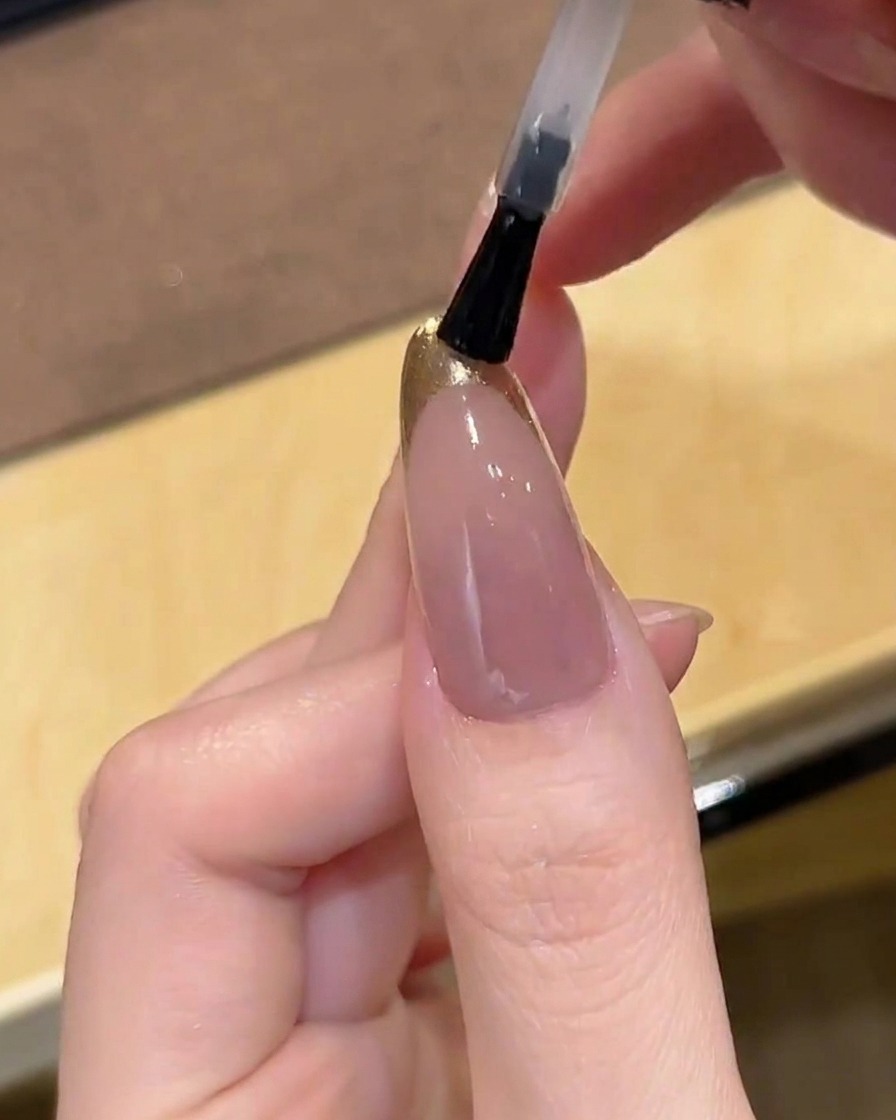 A brush applying a clear, glossy top coat over a gold chrome French-tipped nail.