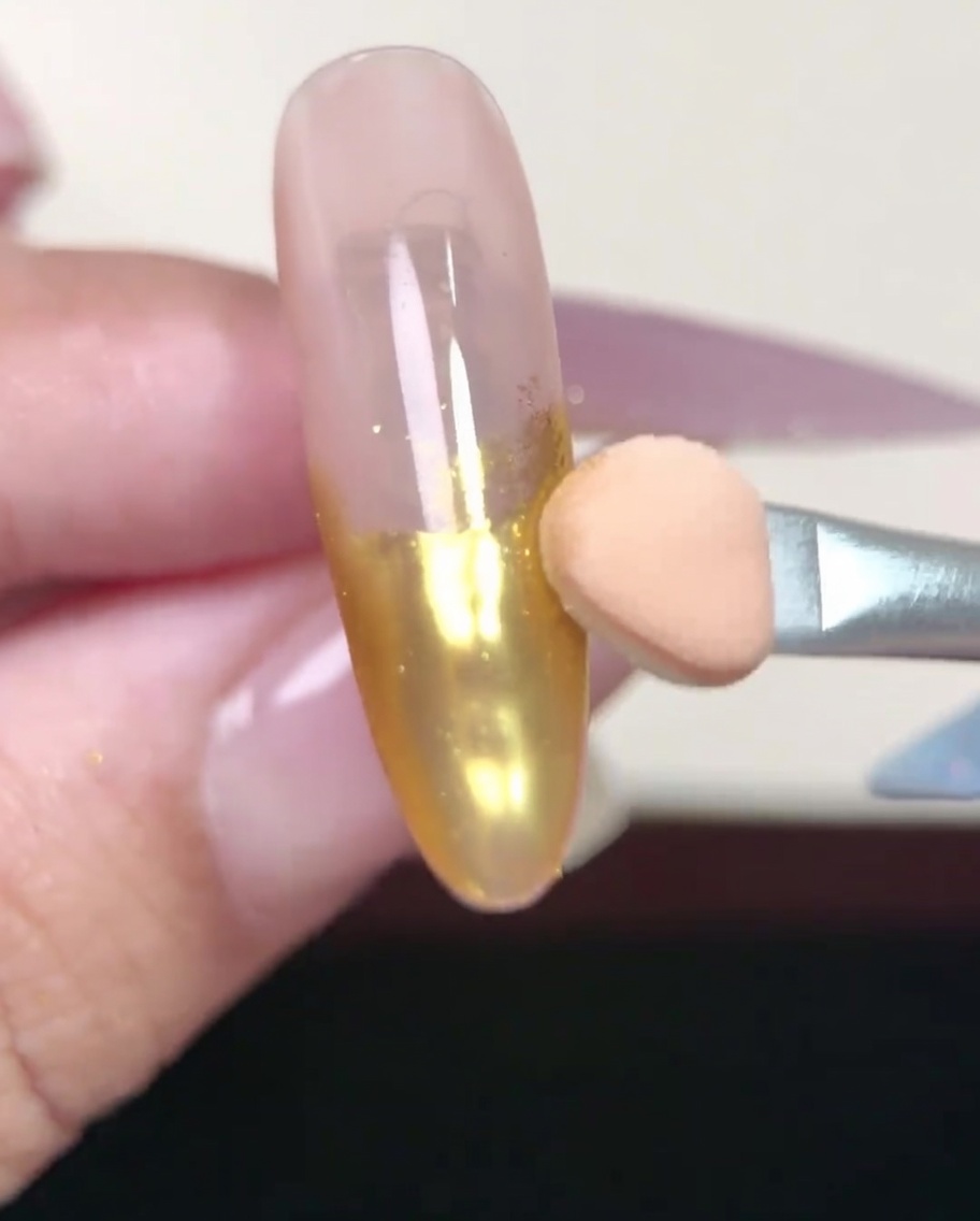 A nail brush smoothly applying a layer of nude gel base coat to a prepped almond-shaped nail.