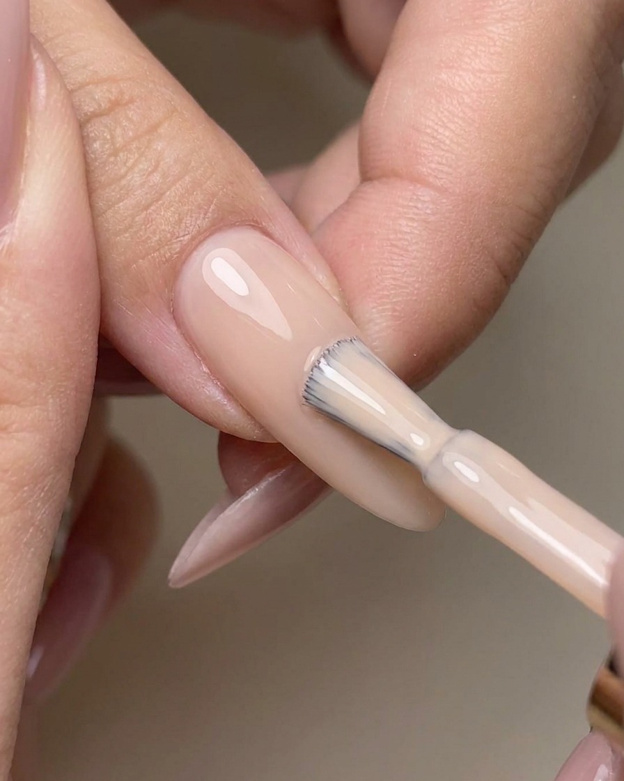 A brush smoothly applying clear no-wipe top coat over a polished nude gel nail.