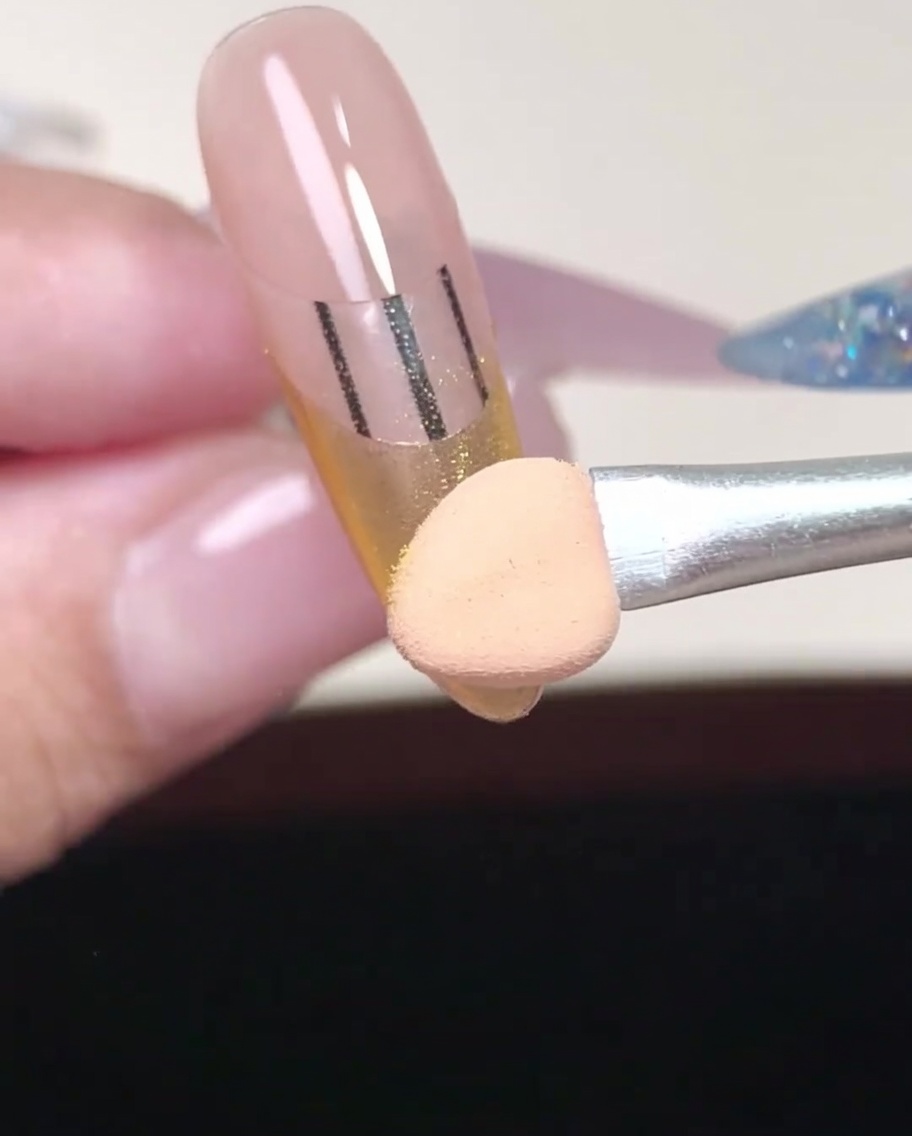Close-up of an applicator tip working gold powder into the corner of a French tip area.