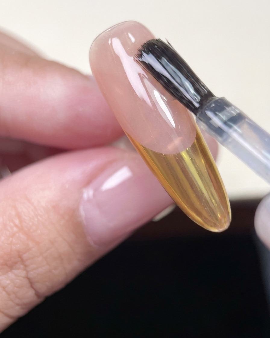 A nail brush applying a clear, glossy top coat over an almond-shaped nail with a gold chrome French tip.