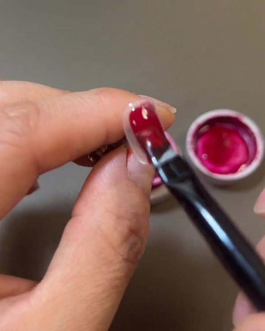 A close-up of a nail brush painting a rich, deep red gel polish onto the nail surface.