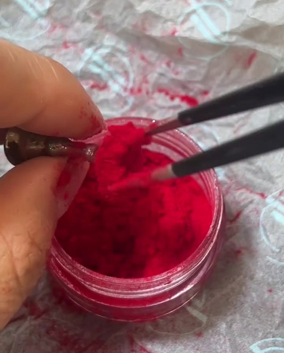 Metal tweezers held over a jar of vibrant red velvet flocking powder, picking up a clump of the fuzzy fibers.