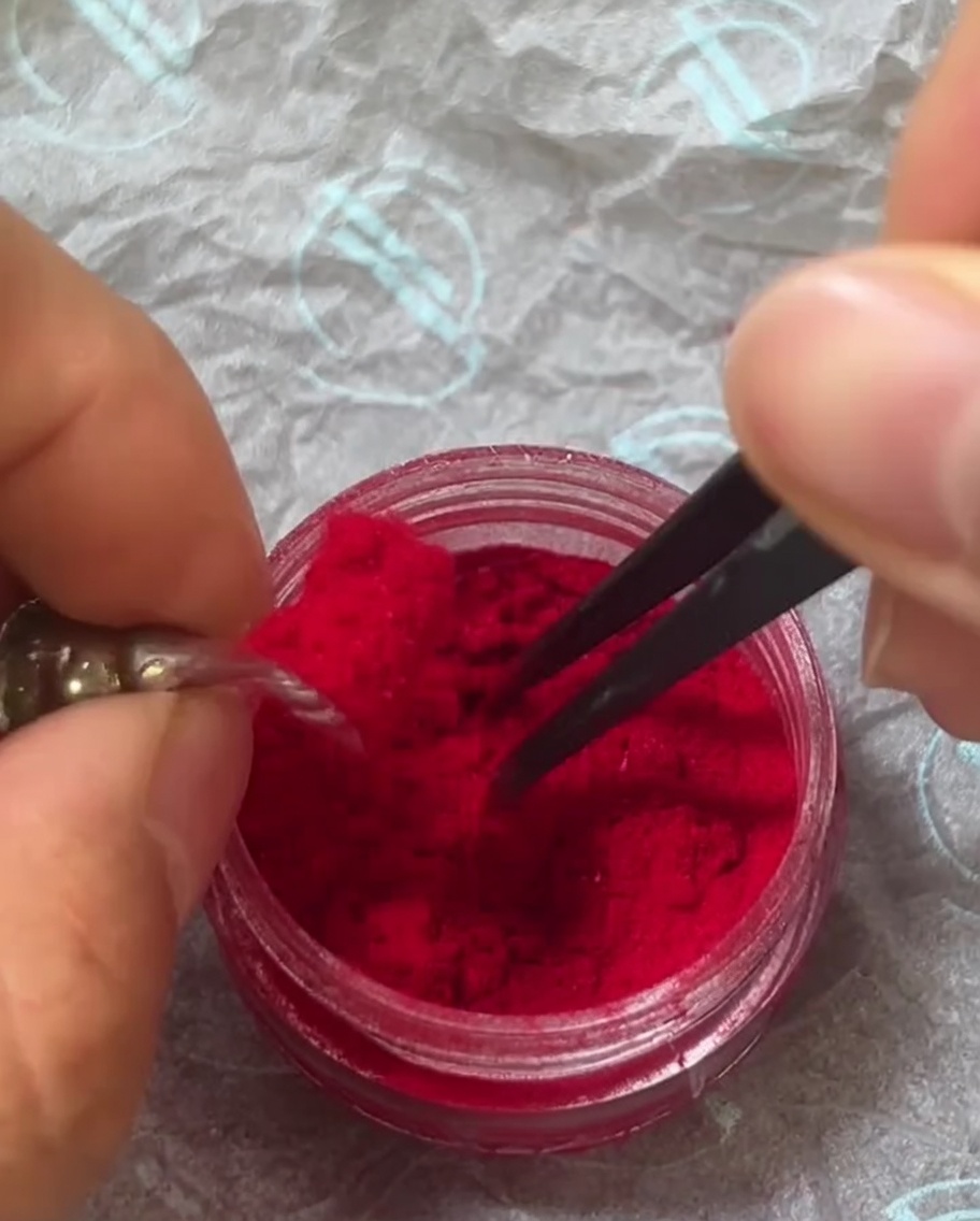 Tweezers carefully sprinkling red fuzzy powder over the surface of a nail that is coated in wet top gel.