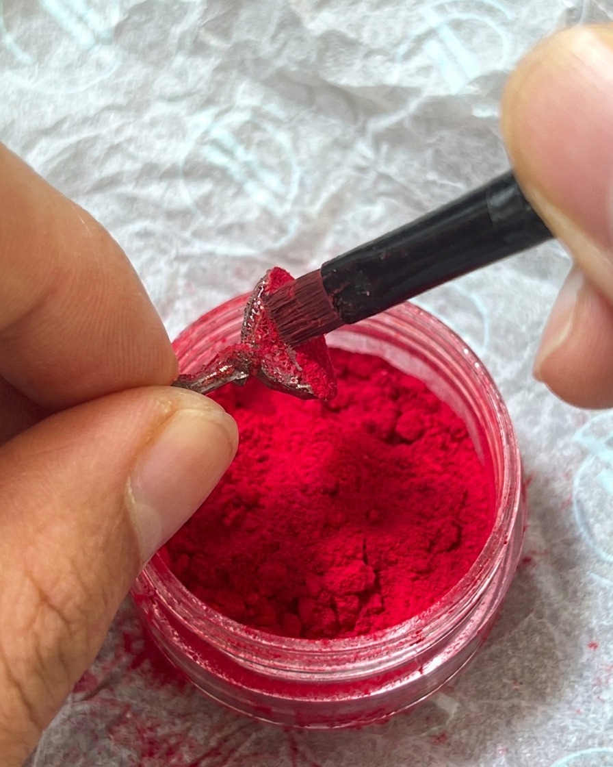 A finger gently pressing down on the red fuzzy powder layered over the nail to secure it into the wet gel.