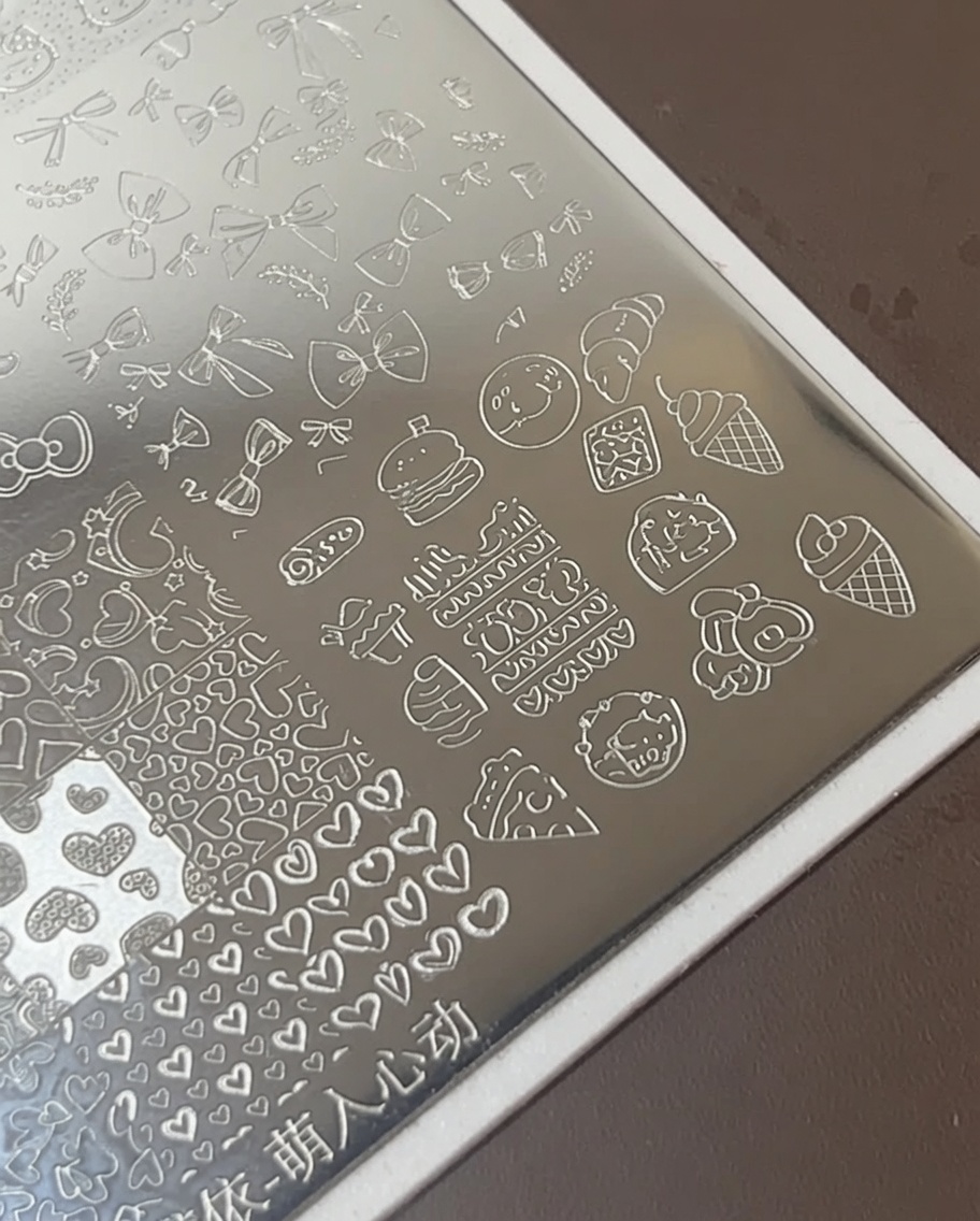 A nail art brush applying thick white polish over etched food icons on a metal stamping plate.