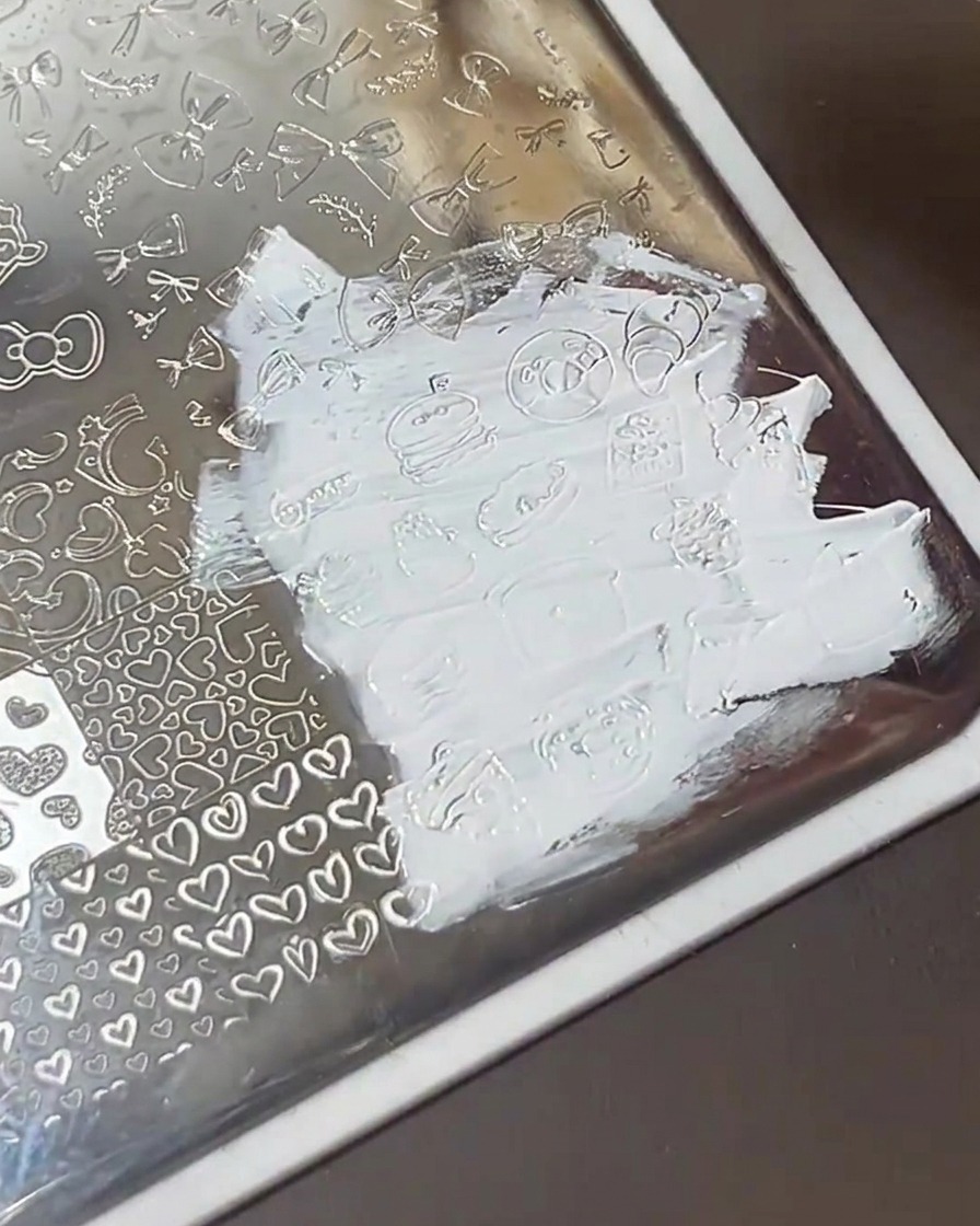 A clear plastic scraper card swiping across a metal stamping plate, leaving clean designs filled with white polish.