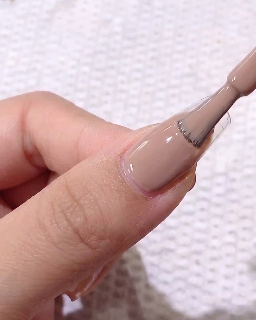 A close-up view of a brush applying a light brown, creamy gel polish to a prepared, clear-coated natural fingernail.
