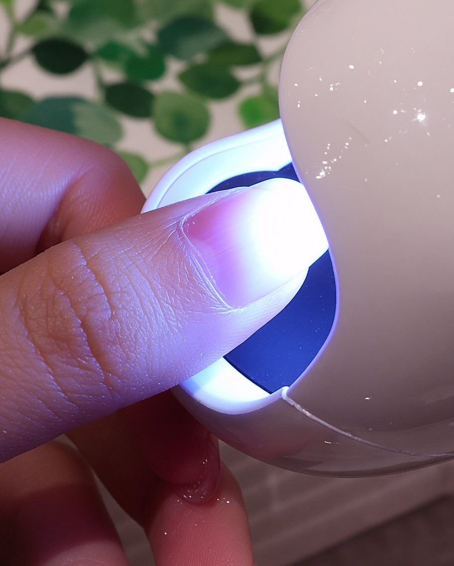 A fingernail with a textured white coating positioned under the purple light of a small UV/LED nail lamp.